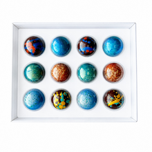Load image into Gallery viewer, A 12-piece set of colorful chocolates arranged in a white square tray. The chocolates feature various patterns including blue, teal, orange, yellow, and black marbling.
