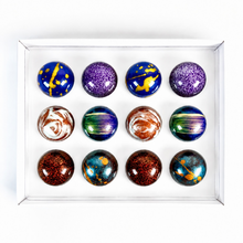 Load image into Gallery viewer, A set of 12 decorative chocolates arranged in a 3x4 grid inside a clear square display box. The marbles feature various colorful patterns including swirls, splatters, and metallic glitter in shades of blue, purple, green, brown, orange, and gold.

